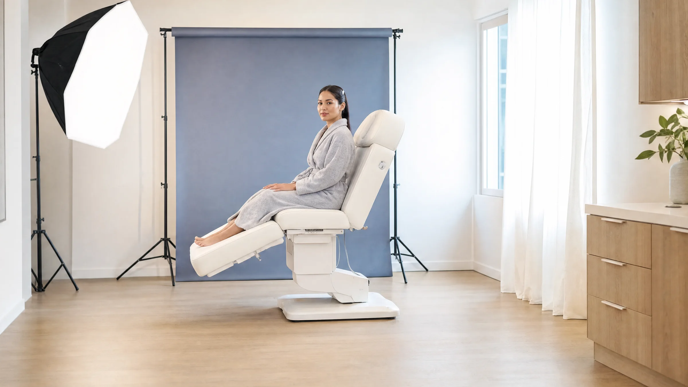 A bright medical aesthetics treatment room with a patient seated in a white chair in front of a blue-gray photography backdrop